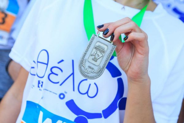 Μια συμμετέχουσα -φορώντας το μπλουζάκι τού εθελοντισμού, με το σήμα «εθέλω»- επιδεικνύει το μετάλλό της