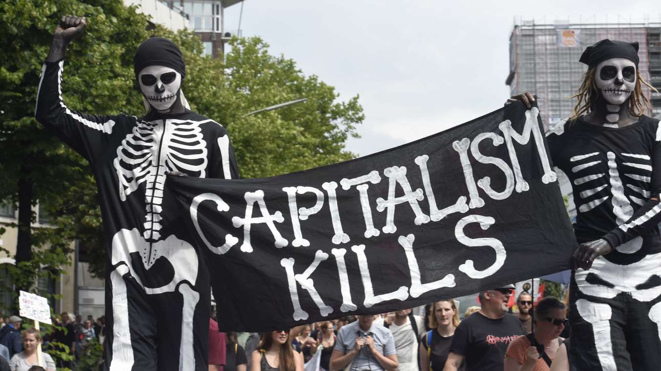 «Ο καπιταλισμός σκοτώνει», γράφει το πανό που κρατούν οι δύο μασκαρεμένοι διαδηλωτές.