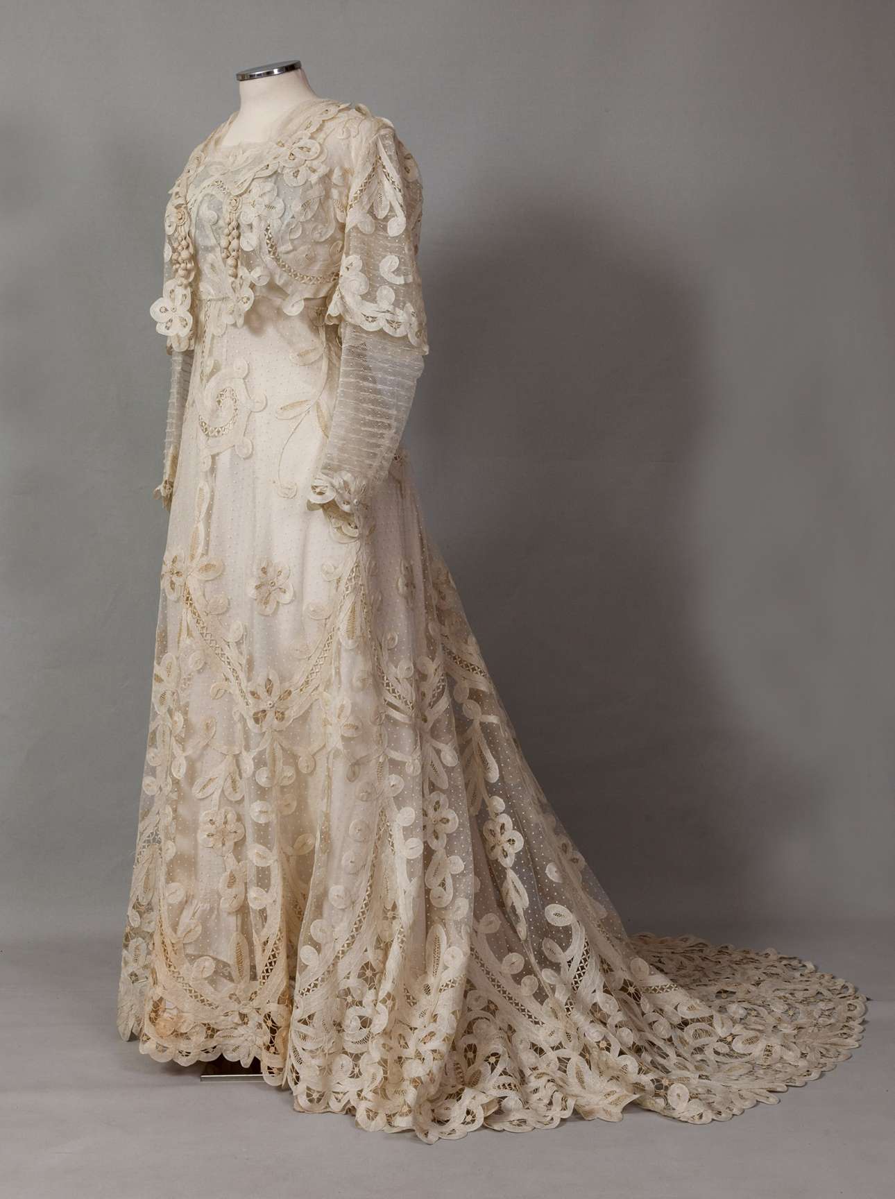 Νυφικό φόρεμα με ουρά από υπόλευκο τούλι και δαντέλα. Φορέθηκε από την Όλγα Κούρτελη στον γάμο της με τον Γιώργο Αντωνιάδη στην Κωνσταντινούπολη το 1898. Δωρεά: Έλλη Σολομωνίδου-Μπαλάνου (Συλλογή ΠΛΙ)