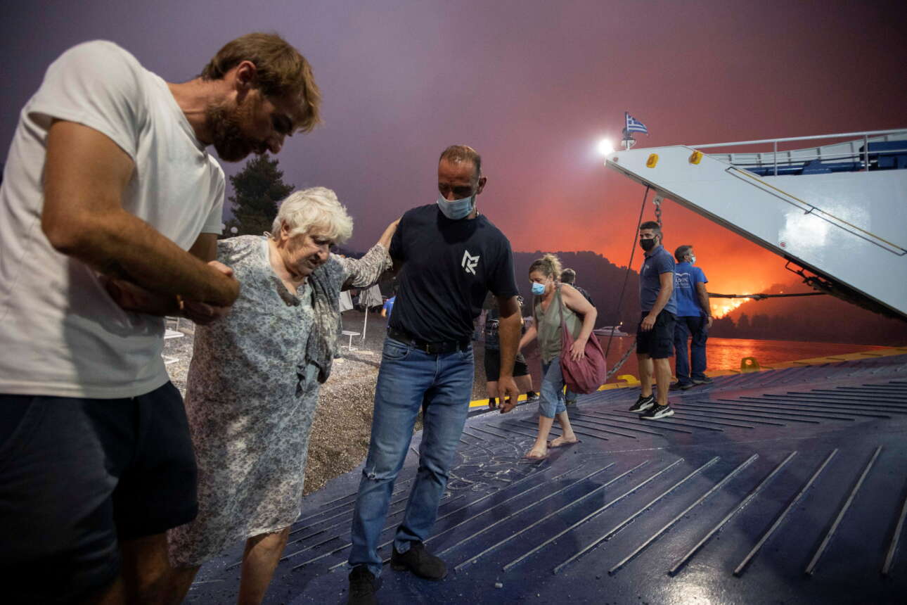 Μια ηλικιωμένη επιβιβάζεται με κόπο και τη βοήθεια των εργαζόμενων στο πλοίο - πίσω οι φλόγες πλησιάζουν