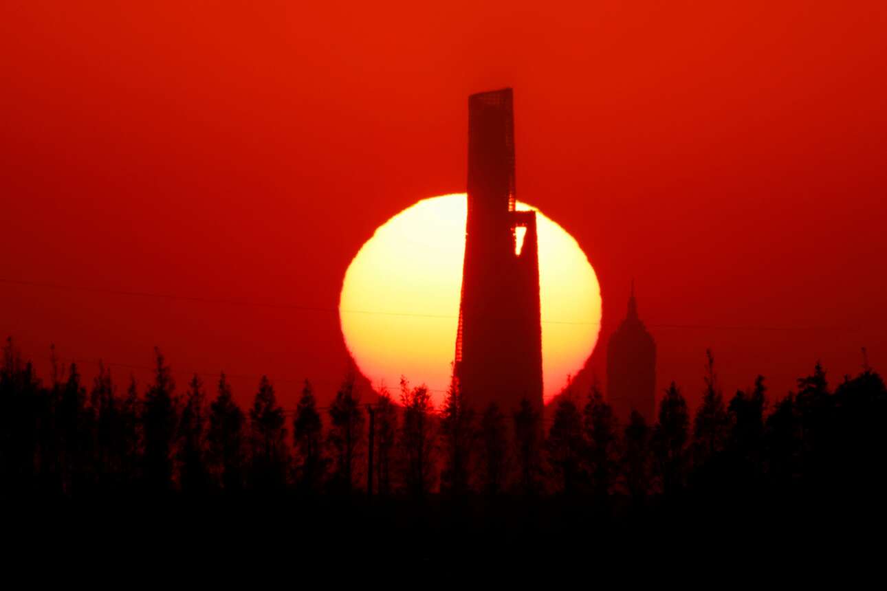 Το ηλιοβασίλεμα βάφει κόκκινο τον ουρανό της Σαγκάης: τα «παλούκια» που του τρυπούν τα σωθικά και κάνουν τα δέντρα να φαντάζουν νάνοι μπροστά τους είναι οι ουρανοξύστες «Πύργος του Μάο» και «Παγκόσμιο Οικονομικό Κέντρο» – κτίρια συμβολικά της «εφόδου» του προέδρου Σι «στον ουρανό»…