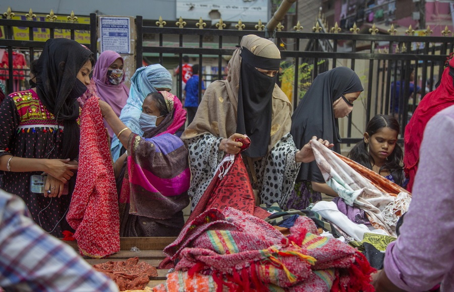 Λαϊκή αγορά στο Μπαγκλαντές, με χειμερινές κολεξιόν: οι γυναίκες, με πρόσωπα κρυμμένα πίσω από υγειονομικές μάσκες και άλλα πανιά, έσπευσαν να αγοράσουν πολύχρωμα τσίτια. Ενα κοριτσάκι έχει σοβαρό ύφος και μία κοπέλα έξυπνο κινητό (με ακουστικά κιόλας)