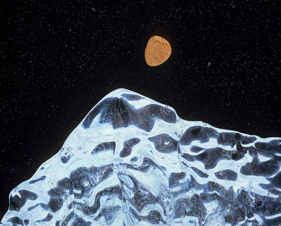Κοιτάζοντας προς τα κάτω ένα παγάκι σε μια παραλία με μαύρη άμμο δίπλα σε ένα στρογγυλεμένο πορτοκαλί βότσαλο, δημιουργείται μια σκηνή που μοιάζει με το φεγγάρι πάνω από μια βουνοκορφή τη νύχτα. Νικητής, Φωτογραφία της Χρονιάς  