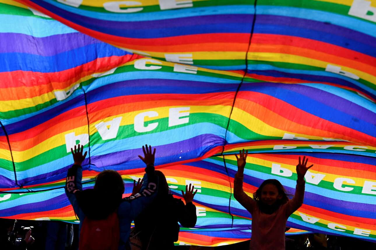 Παιδάκια παίζουν σκεπασμένα από σημαίες με το ουράνιο τόξο στις οποίες αναγράφεται στα Ιταλικά και με κεφαλαία γράμματα η λέξη «ειρήνη» – καρέ από αντιπολεμική διαδήλωση που έγινε στο Μιλάνο 