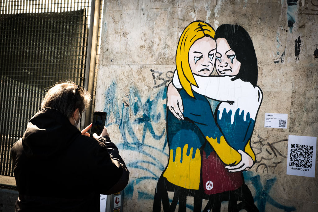 Δύο γυναίκες ντυμένες με τα εθνικά χρώματα της Ουκρανίας και της Ρωσίας αγκαλιάζονται δακρυσμένες στην τοιχογραφία του καλλιτέχνη δρόμου «Laika», στη Ρώμη